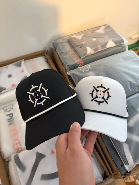 PINSEEKER Performance Golf Caps in Blackout and Whiteout, held over the packing box