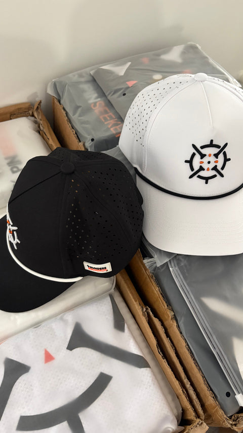 PINSEEKER Performance Golf Caps in Blackout and Whiteout, held over the packing box
