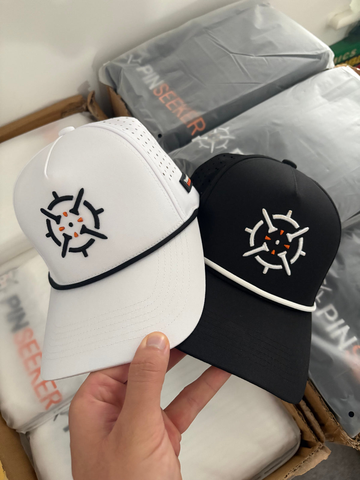 PINSEEKER Performance Golf Caps in Blackout and Whiteout, held over the packing box