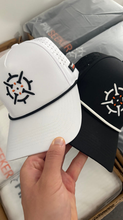 PINSEEKER Performance Golf Caps in Blackout and Whiteout, held over the packing box